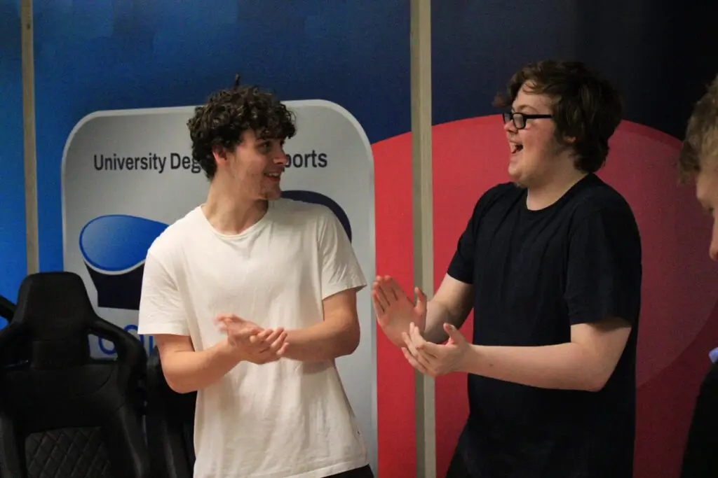 Image depicts two students stood up smiling and clapping together next to a wall with the University Campus of Esports logo on.